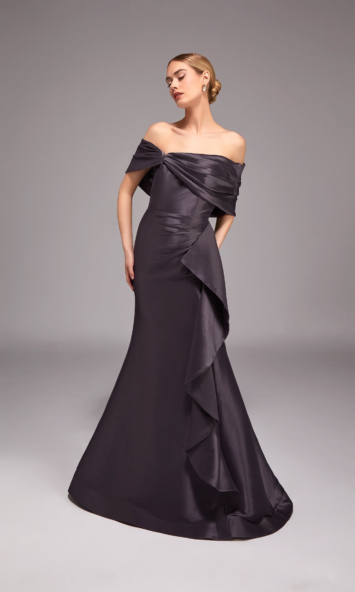 A woman stands against a plain gray background wearing the Alyce 27822 long formal dress in charcoal, featuring an off-the-shoulder neckline, floor-length mermaid silhouette, and ruffled details.